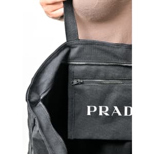 A black tote bag with an interior pocket and a visible logo. The bag's straps are being held by a hand.