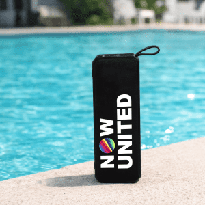 A black portable speaker with a round logo, sitting by a blue pool.
