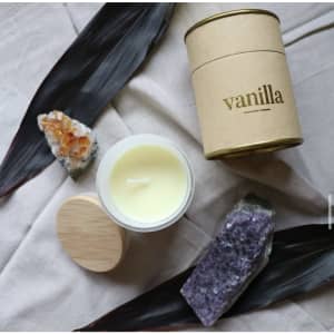 A small vanilla candle in a glass container with a bamboo lid, surrounded by decorative stones and leaves.