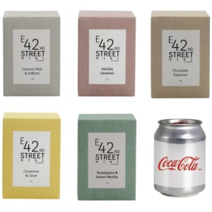 A set of six square candle boxes in various pastel colours: coconut milk and saffron, vanilla caramel, chocolate espresso, cinnamon and clove, eucalyptus and lemon myrtle. A Coca-Cola can is beside them.