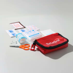 A red first aid kit with scissors, adhesive dressings, antiseptic wipes, and a roll of gauze. The kit includes a logo.