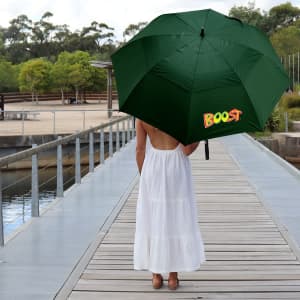 A green umbrella with a logo, held by a person wearing a white dress on a wooden boardwalk near water.