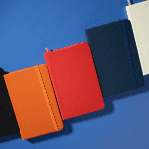 A collection of Minichroma A6 Notebooks in black, orange, red, navy, and white, arranged on a blue surface.