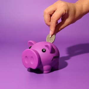 A purple pig-shaped money box with a hand inserting a coin. The background is a solid purple.