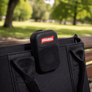 A black portable speaker with a logo, attached to a bag, set against a green park background.