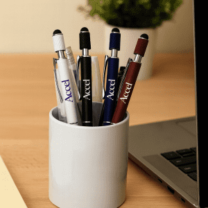 A pen holder featuring several metal pens in white, black, blue, and maroon, placed near a laptop and plant.