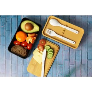 A two-layer lunch box in black and beige contains utensils and food items like fruit, bread, cheese, and vegetables.