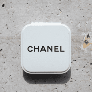 A compact mirror in a white case with a logo on the top, set against a textured stone background.