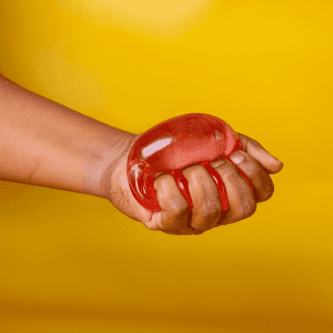 A hand squeezing a translucent red jelly cube sensory toy against a bright yellow background.