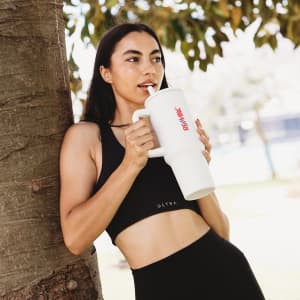 A drink bottle with a large white body and a straw, held by a person in a black sports outfit, leaning against a tree.