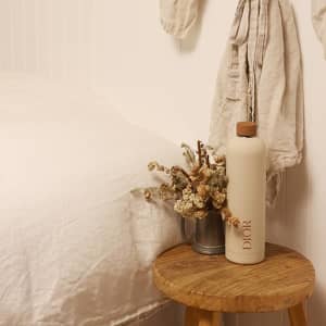 A drink bottle in a soft beige colour with a wooden lid, featuring a logo, sits on a wooden stool beside dried flowers.