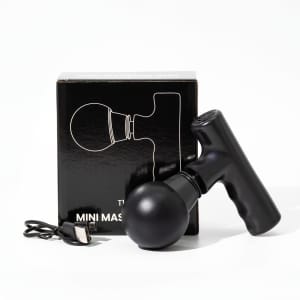 A black mini massage gun beside its box and a charging cable, featuring a sleek design and rounded head.