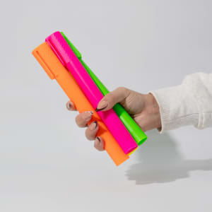 A hand holding three jumbo highlighters in vibrant neon pink, green, and orange colours, featuring a logo.