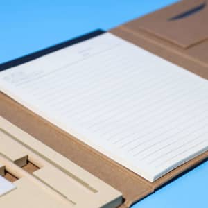 An A5 notepad with lined pages, partially opened within a brown folder. The folder has a logo.
