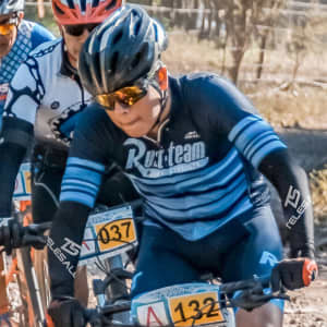 Three cyclists in helmets and sports attire, riding bikes on a dirt path. One is wearing a blue and black jersey.