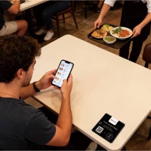 A black NFC epoxy desk tag rests on a table, alongside a customer checking a food ordering app on their phone.