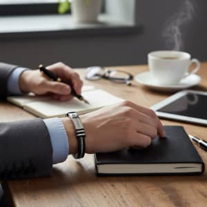 A silver bracelet with a buckle latch rests on a table alongside a notebook, pen, and a cup of coffee.