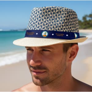 A straw woven top hat with a patterned design and a blue band featuring a logo, set against a beach background.