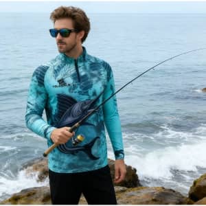 A man wears a blue and teal sublimated 1/4 zip fishing pullover, holding a fishing rod by the sea. It has a logo.