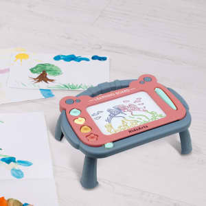 A magnetic drawing board with a stand, featuring grey and pink colours, sits on a light wooden surface beside colourful drawings.