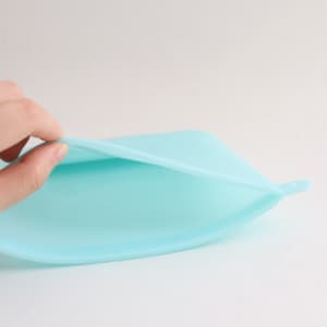 A silicone reusable storage pouch in a light blue colour, with a hand holding it open. It has a smooth surface.