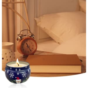 A blue tin candle with a holiday design, surrounded by a bedside table with a clock and a book.
