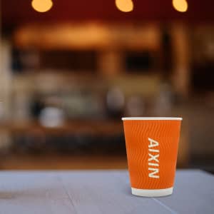 An orange disposable corrugated double wall paper cup with a logo, set against a blurred café background.