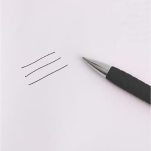 A black barrel ball pen lies beside three lines drawn on a sheet of paper. The pen features a silver tip and grip.