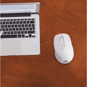 A white wireless mouse placed next to a silver laptop on a wooden desk. The laptop has a visible keyboard.