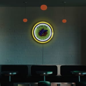 A neon wall clock with a double ring design, featuring a colourful illustration and a logo, set against a brick wall.