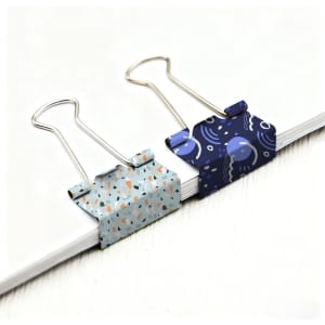 Two decorative binder clips in light blue and dark blue patterns sitting on a stack of white paper.