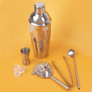 A cocktail shaker set in silver, including a shaker, jigger, strainer, tongs, and ladle on a yellow background.