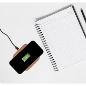 A black smartphone on a square cork wireless charger, placed next to a blank notebook and a pen.
