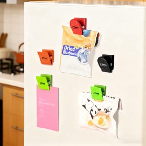 A set of metal magnetic clips in green, orange, black, and red attached to a fridge, holding various items.
