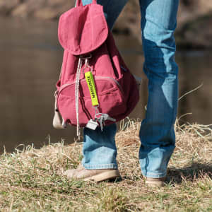 A keyring in yellow attached to a maroon backpack, held by a person standing on grass near a riverbank.