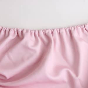 A pink table topper with gathered edges, featuring a smooth fabric texture. It has a branding logo present.
