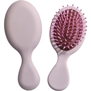 A mini paddle brush in soft pink with a textured grip and flexible bristles. It features a simple logo on the handle.