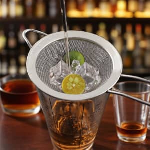 A stainless steel bar mesh strainer with ice and lime slices, positioned over a measuring cup and drinks.