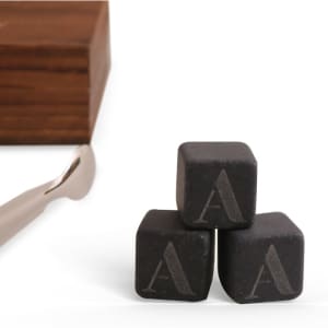 A set of three black whiskey stones with engraved letters, alongside a metal tongs and a wooden box.