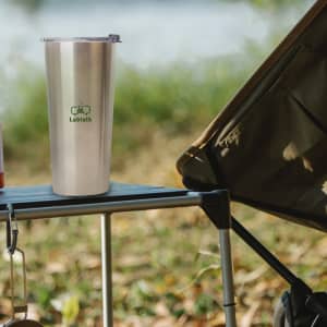 A drink bottle in stainless steel sits on a campsite table, with a logo on its side and a blurred background of nature.