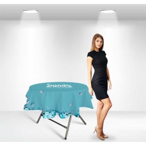A round table cover in turquoise with a floral design, featuring a logo, displayed next to a model in a black dress.