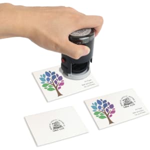 A self-inking stamp in black is being pressed onto white cards featuring a colourful tree design and a logo.