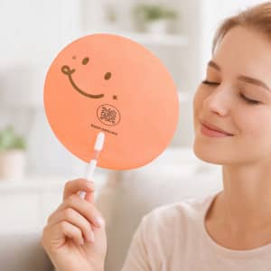 An orange transparent PVC hand fan with a smiling face design and a white handle. Features a logo on the surface.