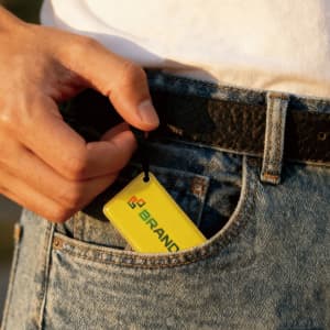 A hand holding a yellow NFC epoxy square tag, with a logo, tucked into the pocket of denim jeans.
