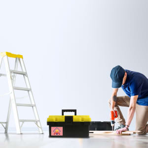 A black tool box with a yellow lid sits on the floor, alongside a person using a drill and a ladder.