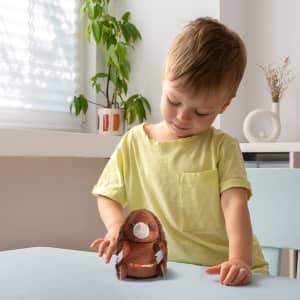 A child in a light yellow shirt holds a brown echidna plush toy on a table, with plants in the background.