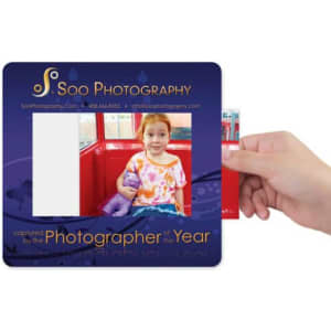 A hand holds a framed mat featuring a colourful design, with an empty space for a photo. It includes a logo.