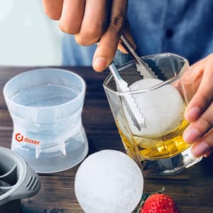 A whisky ice ball mould with a clear sphere of ice, tongs, and a drink in a glass, set on a wooden surface.