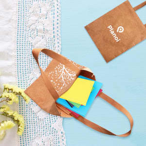 A cork tote bag in brown with a design, containing colourful sticky notes, placed on a light blue surface.