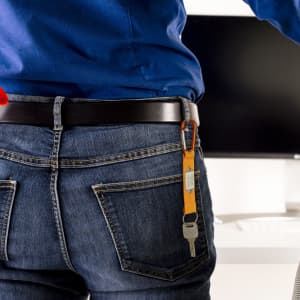 A carabiner keyring in orange attached to a black belt loop, holding keys. A person in blue and denim is visible.
